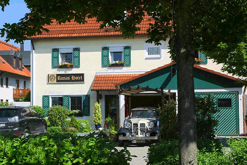 Zweistöckiges, helles Haus mit grünen Fensterläden, Schild Runas Hotel und einem Oldtimer unter einem Vordach, Bäume im Vordergrund.