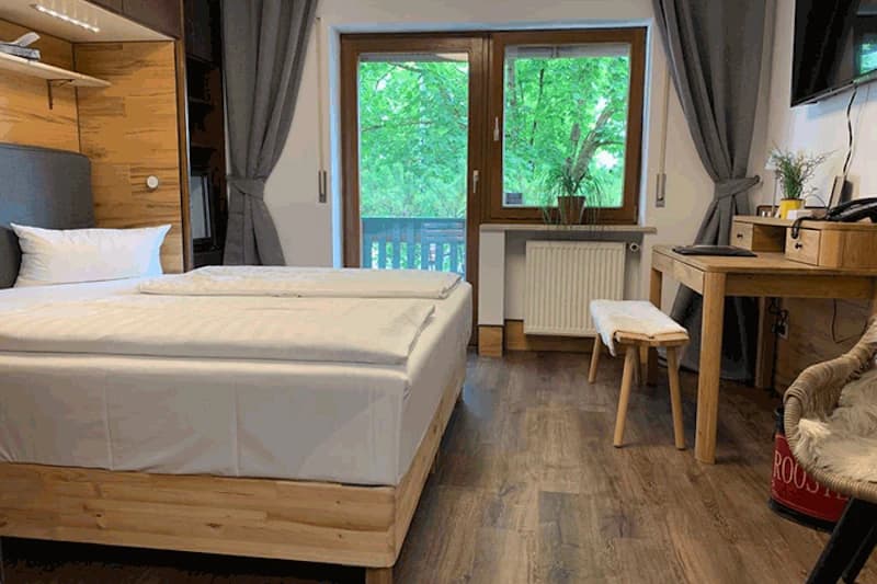Zimmer mit Doppelbett, Holzmöbeln, Schreibtisch, Sitzbank, Balkonfenster und Blick auf grüne Bäume.