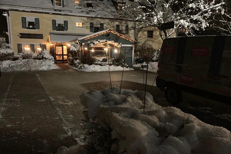 Verschneiter Hof bei Nacht mit beleuchtetem Hoteleingang, geschmücktem Vordach und geparktem Lieferwagen.