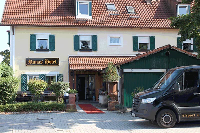Fassadenansicht von Runas Hotel mit grünen Fensterläden, Eingangsüberdachung und schwarzem Bus rechts.
