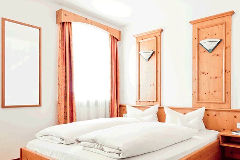 Doppelbett mit weißen Bettdecken, hölzernen Wandpanelen, zwei Wandlampen, Fenster mit Vorhängen.