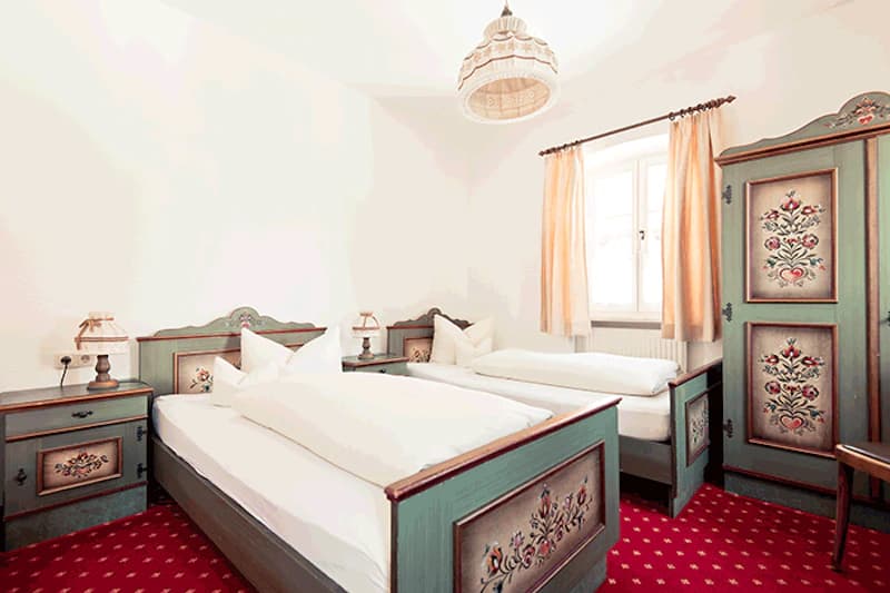Zweibettzimmer mit zwei Einzelbetten, bemalten grünen Holzmöbeln mit Blumenmotiven, rotem Teppich und Fenster mit hellen Vorhängen.