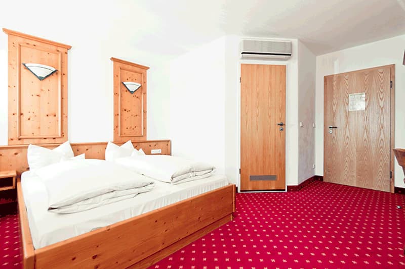 Doppelbett mit Holzrahmen, Kopfteilen und Wandlampen, roter Teppichboden, zwei Holztüren und Klimagerät an weißer Wand.
