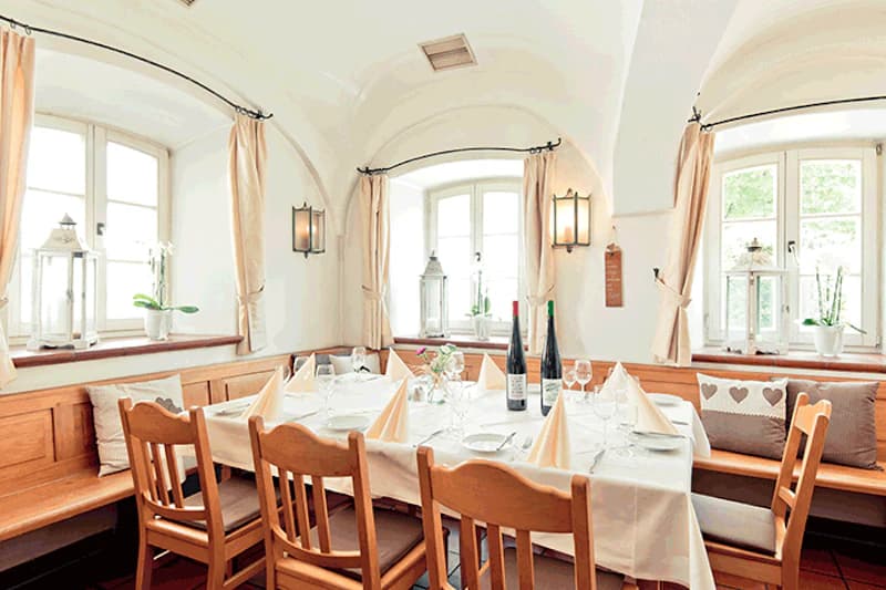 Gedeckter Holztisch in gemütlicher Stube mit Bank, Holzstühlen, weißen Servietten, Weinflaschen und Fenstern mit Vorhängen.