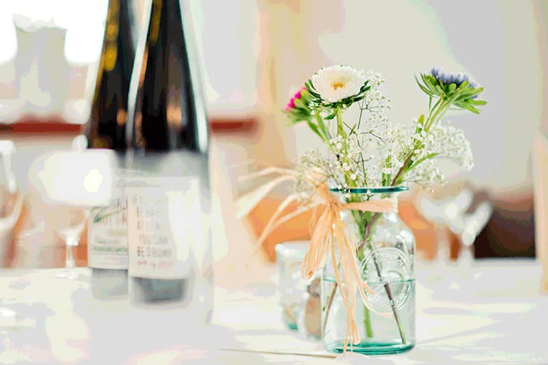 Glasvase mit Schleife und kleinen weißen und rosa Blumen auf gedecktem Tisch, im Hintergrund Weinflaschen unscharf.