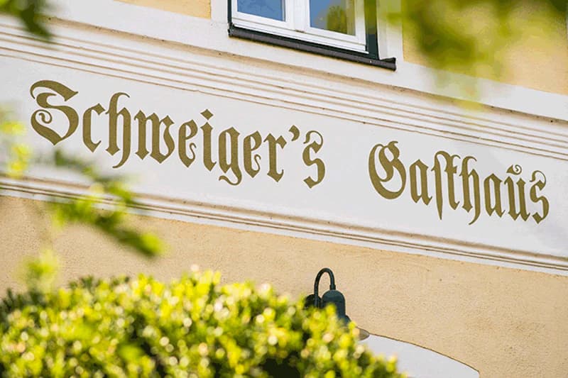 Fassade mit Schriftzug Schweiger's Gasthaus in Frakturschrift, Fenster, Laterne und grüner Bepflanzung.