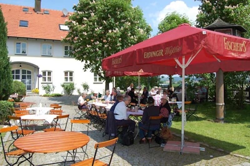 Gartenterrasse mit runden Holztischen, Bierbänken, rotem Erdinger-Sonnenschirm und Gästen vor weißem Haus.