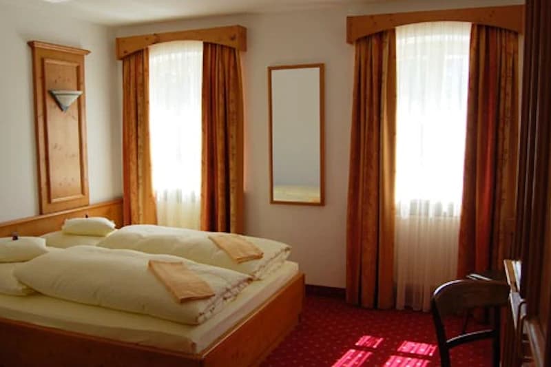 Hotelzimmer mit Doppelbett, zwei Fenstern mit Vorhängen, Spiegel, Holzmöbel und rotem Teppich.