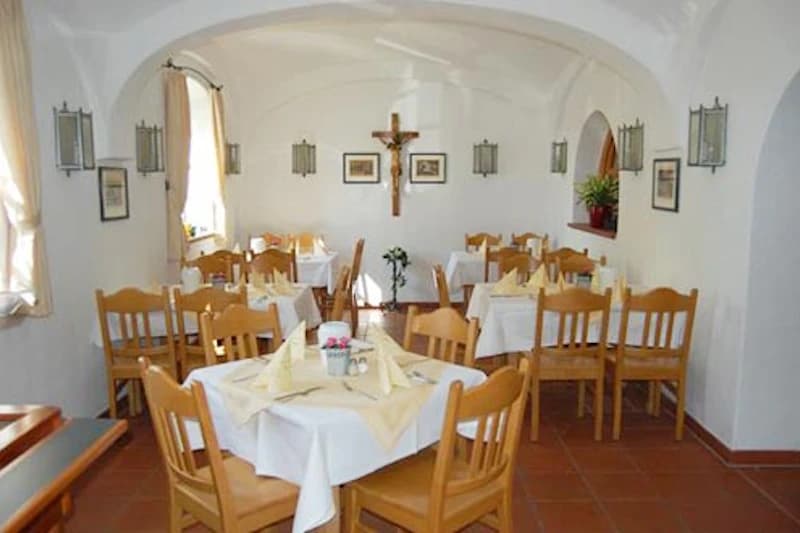 Speiseraum mit Holztischen und -stühlen, weißen Tischdecken, gefalteten Servietten und Kruzifix an der Wand.