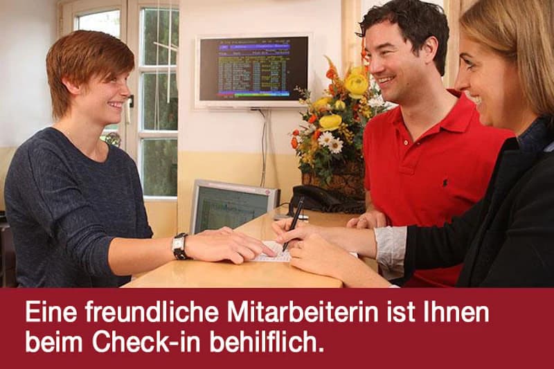 Rezeption mit Mitarbeiterin links, Paar rechts unterschreibt am Tresen, Monitor mit Anzeigetafel im Hintergrund.