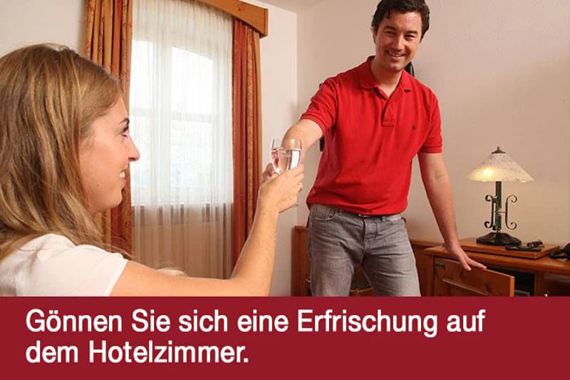 Mann im roten Poloshirt reicht Frau ein Glas Wasser in Hotelzimmer mit Fenster und Lampe.