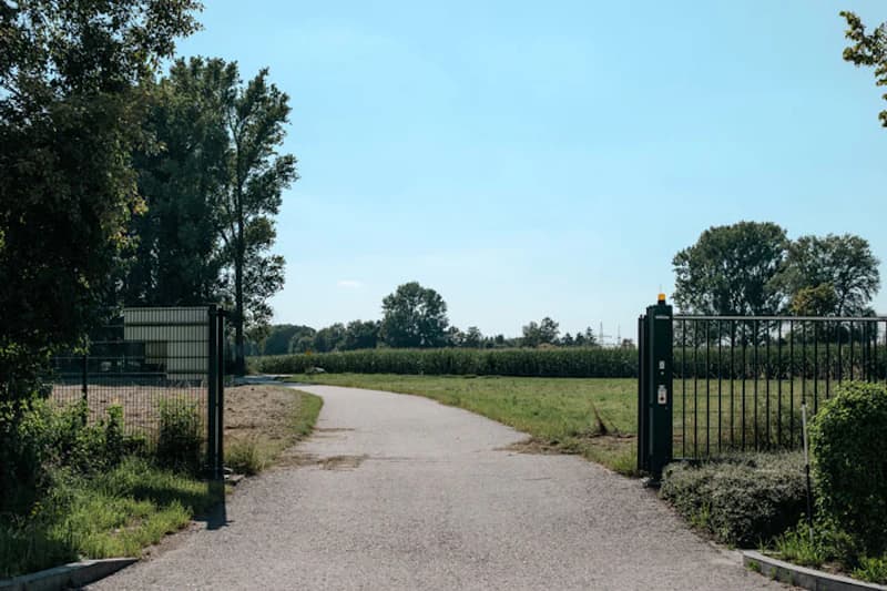 Offenes Metalltor an einer asphaltierten Zufahrt, Zaun links, Steuerpfosten mit Bedienelement rechts, Felder und Bäume im Hintergrund.