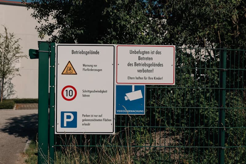 Metallzaun mit Tor und Schildern: Betriebsgelände, Schrittgeschwindigkeit 10, Parkhinweis und Videoüberwachung.