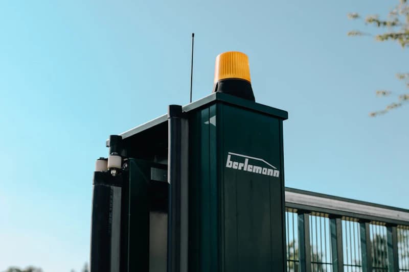 Pfosten einer elektrischen Toranlage mit gelbem Warnblinker und Firmenlogo vor blauem Himmel.